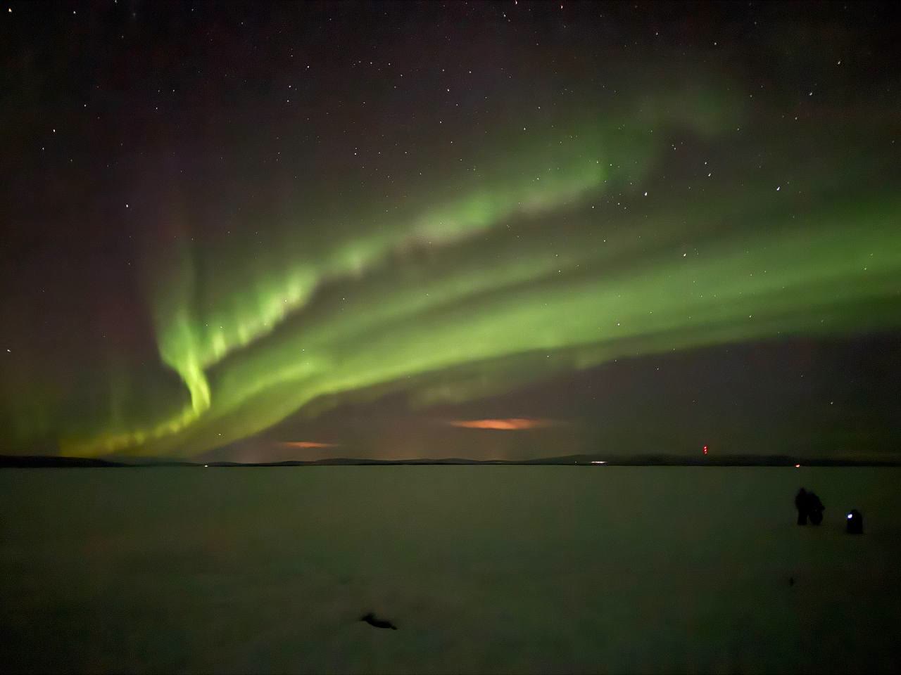 Green arcs of aurora over a frozen lake with people watching